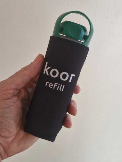 Hand holding a green koor bottle with a black sleeve labeled 'Koor' on a light background